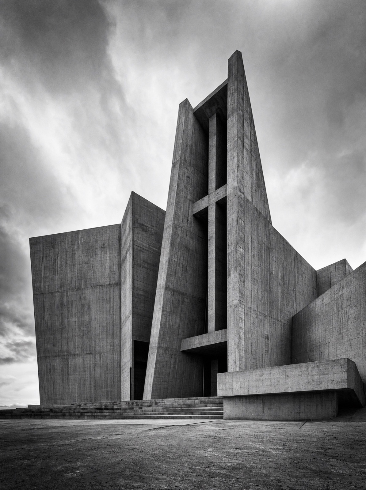 Angular concrete sanctuary with a split bell tower against a stormy sky